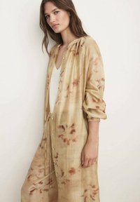 Camicia loose-fitting beige chiaro con motivo floreale, apertura con bottoni, maniche lunghe e texture morbida. Abbinata a una canotta bianca sotto.