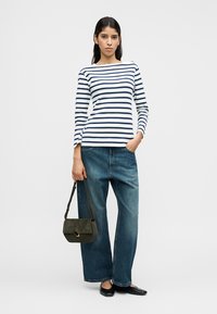 Ριγέ μακρυμάνικη μπλούζα σε λευκό και ναυτικό μπλε, συνδυασμένη με φαρδιά μπλε τζιν. Το μοντέλο κρατά μια σκούρα πράσινη σουέντ τσάντα και φοράει μαύρα φλατ παπούτσια.