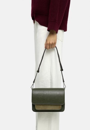 Osoba w bordowym swetrze i białych spodniach trzyma ciemnozieloną skórzaną torebkę z teksturowaną przednią kieszenią przy pasku.