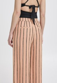 Pfirsichfarbene weit geschnittene Hose mit vertikalen schwarzen Streifen, ausgestattet mit einem hohen Bund und zwei Gesäßtaschen. Hergestellt aus leichtem Stoff.