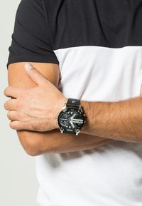 Orologio analogico nero con grande quadrante rotondo, dettagli argentati, funzioni cronografiche e cinturino in pelle nera. Indossato su un polso con pelle chiara.