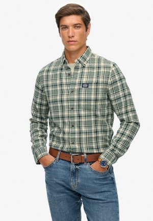 Camisa a cuadros verde y crema con botones, mangas largas, puños con botones y un bolsillo en el pecho. Combinada con jeans de mezclilla azul y un cinturón marrón.