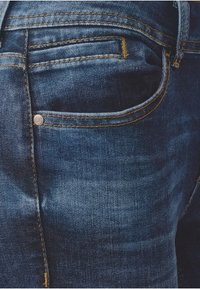 Jeans de mezclilla azul oscuro con un diseño clásico de cinco bolsillos, costuras en contraste y un pequeño detalle de logotipo metálico en el bolsillo trasero.