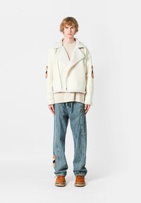 Chaqueta blanca forrada de borrego, con parches bordados, superpuesta a una blusa beige y combinada con pantalones de denim azul sueltos y zapatos marrones.