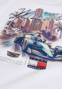 Formel 1 racerbiler, der kører hurtigt på Miami-banen med palmer, byens skyline, ternede flag, Cadillac- og Tommy Hilfiger-logoer samt dato for begivenheden.