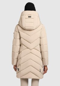 Persoon in een beige gewatteerde pufferjas met capuchon en zwarte gebreide beanie, gezien van achteren tegen een effen achtergrond.