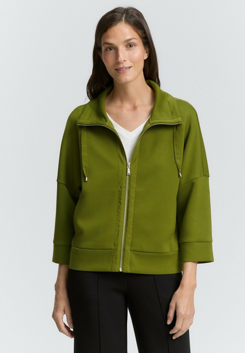 Veste zippée verte avec un col haut, des manches longues et deux poches latérales. Tissu doux avec une texture lisse et des détails de conception simples.