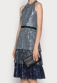 Abito senza maniche con paillettes in tonalità di blu, con strati a balze e cintura nera in vita, abbinato a una pochette nera. Tessuto liscio e finitura lucida.