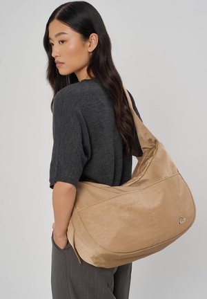 Vrouw met lang donker haar, gekleed in een donkergrijs shirt en grijze broek, draagt een grote beige schoudertas met een klein hartembleem.