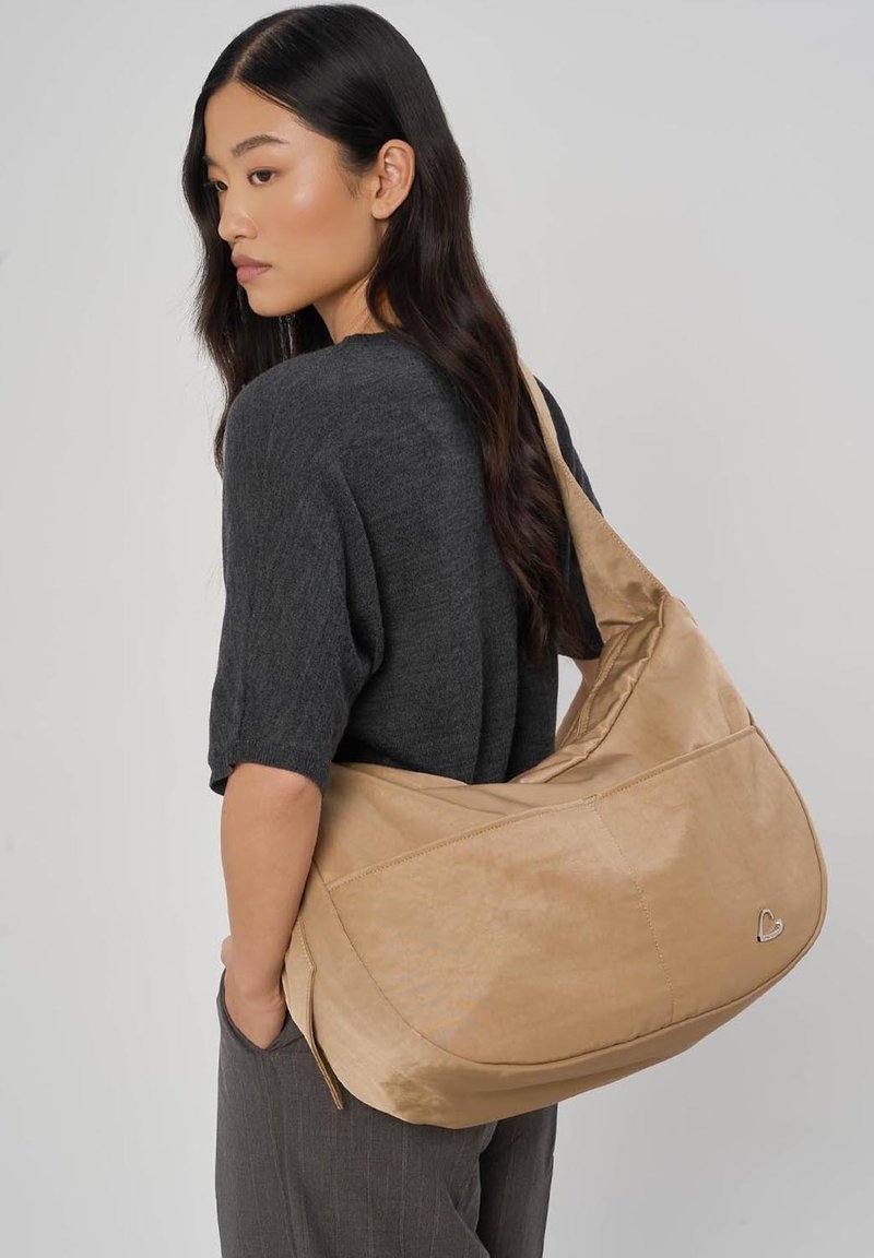 Femme aux longs cheveux noirs portant un haut gris foncé et un pantalon gris, avec un grand sac à bandoulière beige orné d'un petit emblème en forme de cœur.