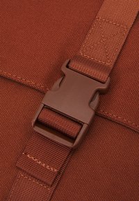 Gros plan sur un sac messager en tissu brun, avec une boucle en plastique et une sangle réglable. Matériau texturé avec des coutures visibles.