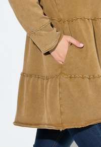 Abito marrone con texture, caratterizzato da un orlo arricciato e maniche lunghe, con tasche laterali e cuciture a vista, abbinato a jeans blu.