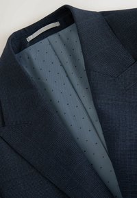 Donker marineblauwe blazer van een gestructureerde stof. De binnenkant heeft een lichtblauwe voering met kleine zwarte stippen. Opvallende gestructureerde schouders.