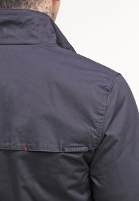 Námořnicky modrá bunda se stojat�ým límcem, hladkou texturou a viditelnou černou značkovou cedulkou na švu. Šití obsahuje malý červený akcent.