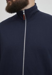 Veste zippée bleu marine en matériau doux avec un col haut, dotée d'une fermeture éclair en argent et d'une tirette en cuir marron.