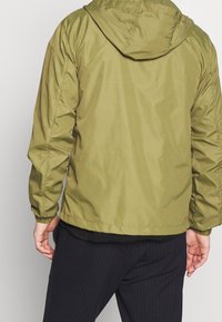 Olivgrön huvvjacka med en glänsande, slät yta, featuring elastiska muddar och en midjelång längd. Enkel design utan synliga logotyper.