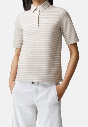 Persona con indosso una polo a maniche corte beige e bianca a righe, infilata parzialmente nei pantaloni bianchi, con le mani nelle tasche, su uno sfondo semplice.