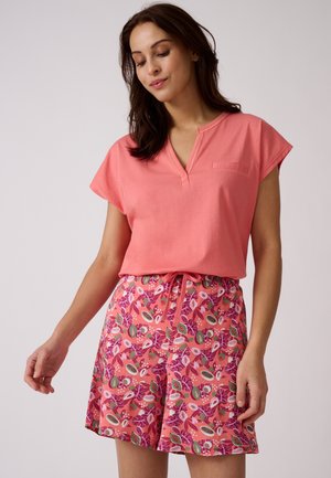 Vrouw draagt een koraalroze top met korte mouwen en roze short met een kleurrijk tropisch fruitpatroon, staat met gesloten ogen.