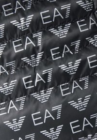 Vadderad tyg med ett upprepande vitt EA7-logotyp och vingdesign på en svart bakgrund. Slät struktur med kviltade sömmar.