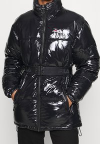 Giacca a vento nera con texture lucida, colletto alto e chiusura con zip. Presenta una cintura nera con il logo FILA e tasche laterali.