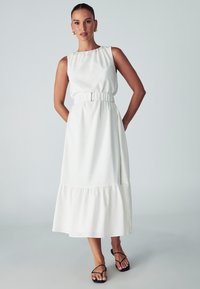 Sleeveless white dress with a gathered waist, flowing hem, and tiered bottom. Worn with black strappy sandals. Smooth fabric texture.