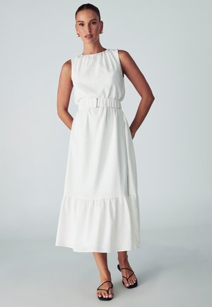 Ärmelloses weißes Kleid mit gerafftem Bund, fließendem Saum und gestuften Boden. Tragen mit schwarzen Riemchensandalen. Glatte Stofftextur.