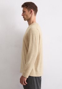 Pull beige à manches longues avec un col simple, texture douce et coupe décontractée. Porté avec un pantalon gris foncé, vu de côté.