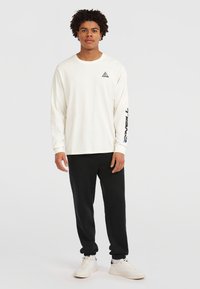 Maglietta bianca a maniche lunghe con un piccolo logo triangolare sul petto, pantaloni neri, sneakers bianche e dettagli neri sulle maniche.