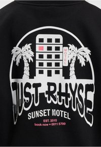 Fekete kapucnis pulóver grafikai tervezéssel, amelyen egy szállodaépület, pálmafák, valamint a „JUST RHYSE” és a „SUNSET MOTEL” felirat fehér és rózsaszín színben található.