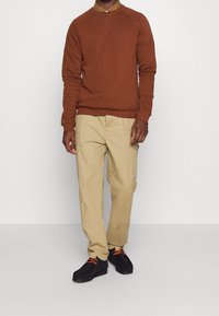 Felpa marrone con maniche raglan, abbinata a pantaloni cargo beige con tasche laterali e scarpe nere con lacci arancioni.
