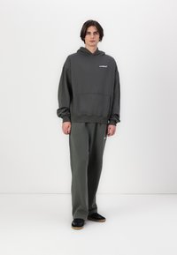 Dark gray hoodie with a front pocket and logo, paired with matching sweatpants. Model stands, wearing black shoes with a gum sole.