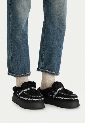 Piernas con vaqueros azules remangados, calcetines de punto beige y mocasines negros sin cordones con costuras blancas en zigzag y forro de piel sintética.