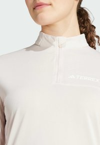 Lichtroze Terrex lange mouwen top met een ritssluiting kraag, zachte textuur en een minimalistisch design met een klein wit logo op de borst.