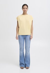 T-shirt gialla a maniche corte abbinata a jeans svasati azzurri chiari e tacchi neri, su uno sfondo bianco uniforme.