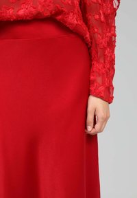 Robe rouge avec un haut transparent à manches longues texturé, orné de broderies florales. La jupe est lisse et ajustée, assortie à la couleur vive du haut.