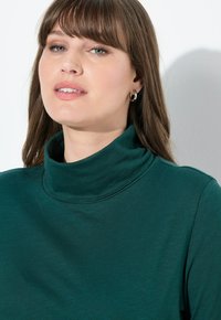 Maglione a collo alto color turchese realizzato in tessuto morbido, con un collo aderente e una superficie liscia. Design minimalista senza motivi visibili.