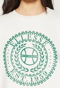 Felpa bianca con un logo circolare verde raffigurante un pallone da basket, foglie di alloro e la scritta "ELLESSE EST. 1959" in un carattere bold.