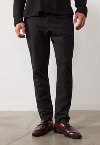 Pantalon noir sur mesure en tissu lisse, associé à des mocassins bordeaux brillants avec deux brides, sur un sol en béton.