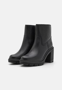 Botas negras de tobillo de cuero con puntera cuadrada, tacón grueso y suela de goma texturizada. Presentan un cierre de cremallera lateral y costuras sutiles.