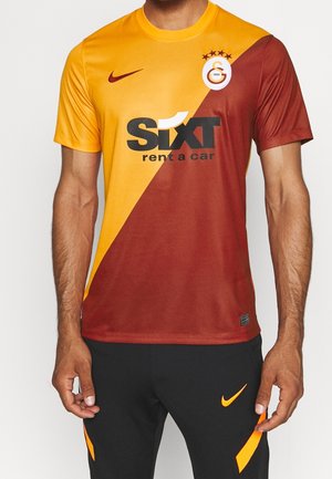 Mężczyzna noszący żółto-czerwoną koszulkę piłkarską Galatasaray w stylu połowa-połowa z logo Sixt rent a car i czarne spodnie Nike.