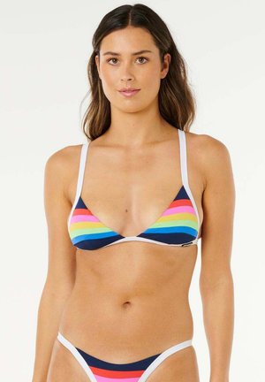 Femme portant un bikini bleu marine avec des rayures arc-en-ciel horizontales et des bretelles blanches, debout devant un fond clair uni.