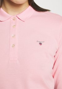 Rosa polotröja med krage, som har fem knappar och en liten broderad logotyp på vänster bröst. Texturerad bomullstyg.