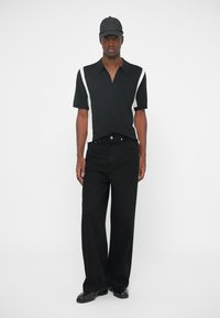 Polo preto de manga curta com detalhes laterais brancos, combinado com jeans pretos de perna larga e sapatos pretos, sobre um fundo simples.