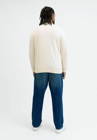 Langermige crèmekleurige trui met geribde manchetten en zoom, gecombineerd met een losse blauwe spijkerbroek en witte sneakers, gezien van achteren.