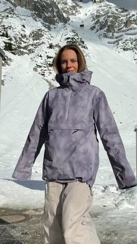 Veste imperméable violette avec capuche, fermeture éclair à l'avant et poignets élastiques, assortie à un pantalon de neige beige clair, le tout sur fond de montagne enneigée.