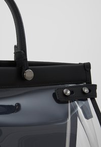 Borsa trasparente con parte superiore e manici in pelle nera. Presenta hardware in tonalità argento, cuciture visibili e due accenti a bottone.