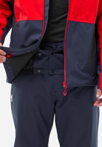 Rote und marineblaue, wasserdichte Jacke mit Reißverschlusstaschen. Passende marineblaue Hose mit Druckknopfverschluss, glatter Textur und verstellbarem Sitz.