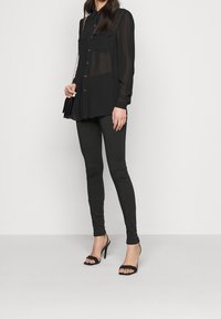 Camisa negra transparente de manga larga con botones en el frente y dos bolsillos en el pecho, combinada con leggings negros ajustados y sandalias de tacón. Incluye un bolso sencillo.