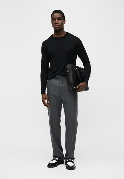 Homme portant une chemise noire à manches longues et un pantalon gris, tenant une mallette en cuir noire, debout sur un fond clair uni.