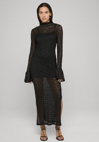 Robe longue noire en dentelle avec manches longues, col haut et manches évasées. Comprend une superposition transparente et des fentes latérales, mettant en valeur la silhouette.